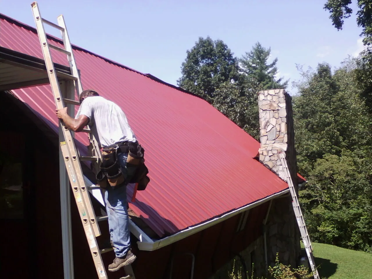 Roofer on a ladder beside a red metal roof and stone chimney — Soffit & Fascia Repair by Roofing Brothers in Kouts