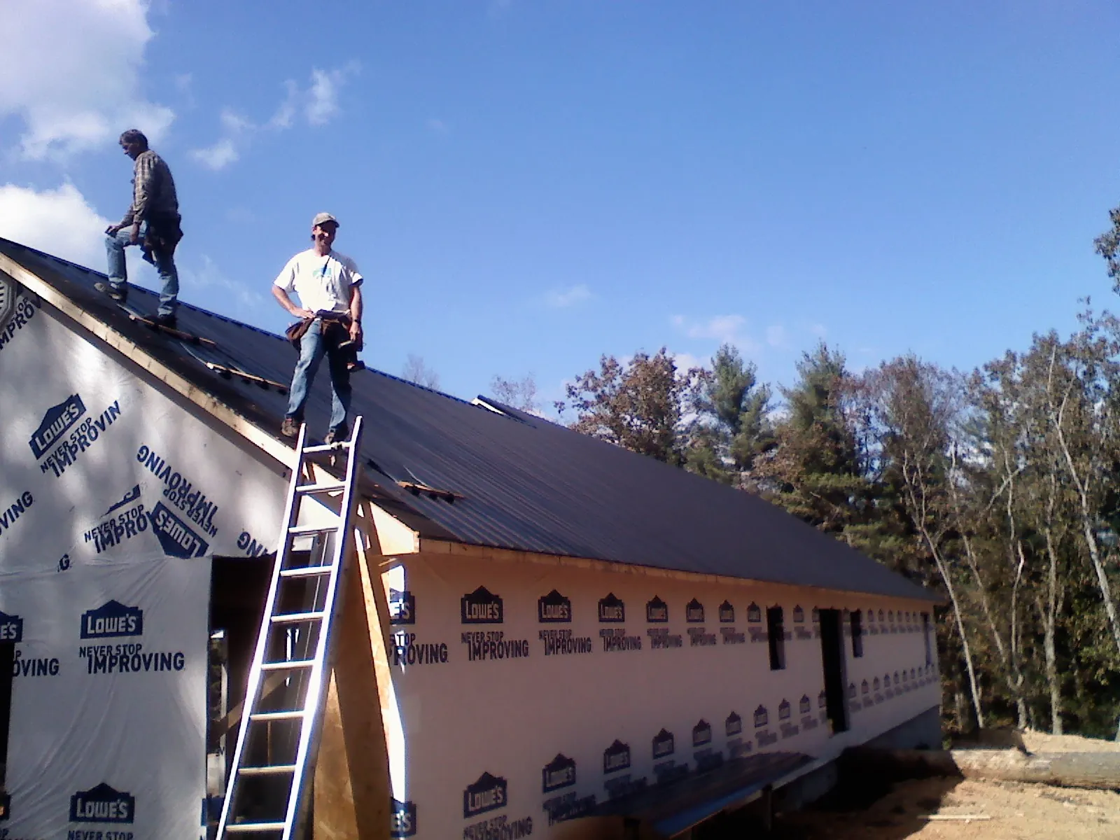 Two roofers standing on a long dark metal roof over a wrapped building in Kouts